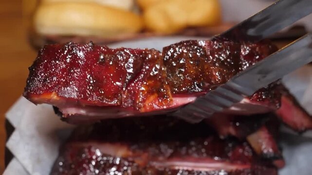 Succulent, glazed pork ribs are presented in a close-up, highlighting their rich color and tender texture. A pair of metal tongs holds a single rib to reveal the smoke ring and juicy interior of the s