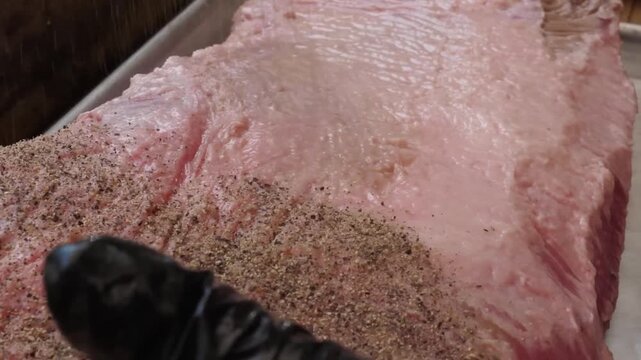 Close-up of a large, fresh raw beef brisket cut, generously seasoned with salt and pepper on a metal baking sheet. This high-quality piece of meat features visible fat rendering and marbling, ready fo