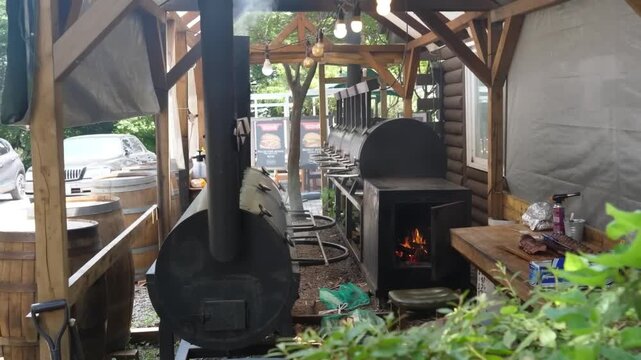 A high-angle shot showing a large, professional-grade black offset smoker trailer in an outdoor covered patio. Glowing embers and flames are visible inside the open firebox door as smoke rises from th