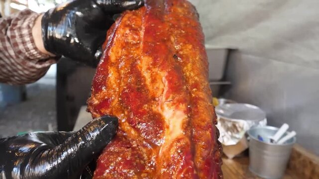 A professional chef or pitmaster, wearing black nitrile gloves, holds up a juicy, glistening rack of slow-smoked pork spare ribs. The meat is generously coated in a vibrant, caramelized red barbecue s