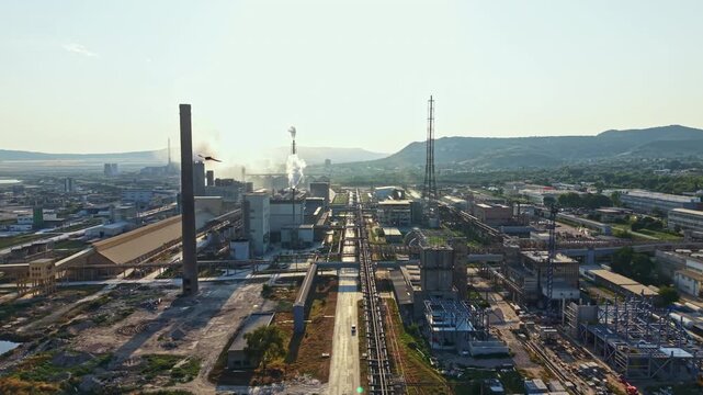 The scene shows an industrial area with multiple factories, smokestacks, and power lines. Smoke rises from the factories under a clear sky. Roads and paths are visible in the foreground.