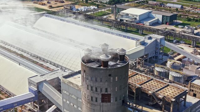 Large industrial plant with machines and buildings stands in a flat area. Smoke rises from stacks as workers operate around the site. It is daytime and the sky is clear.