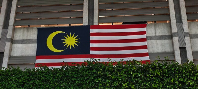 The Jalur Gemilang, Malaysia&rsquo;s national flag, displayed on the exterior of a modern building with horizontal slats and greenery below. 