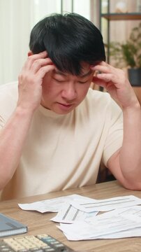 Asian man sitting at home table grips head, unpaid bills scattered with calculator and laptop, shoulders droop. Chinese guy sighs, showing financial stress and urgently seeking workable answer now.