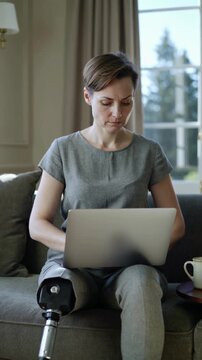 Woman with prosthetic leg sitting on sofa typing on laptop computer, medium shot