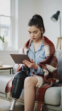 Young woman with prosthetic leg using tablet on sofa with red plaid blanket, medium shot