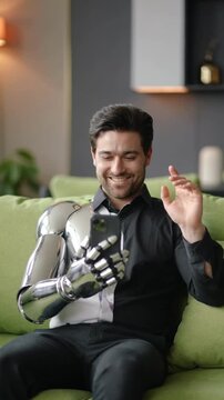 Man with metallic prosthetic arm video calling on smartphone sitting on green sofa medium shot