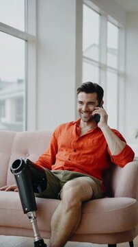 Young man with prosthetic leg sitting on sofa talking on mobile phone in bright modern living room