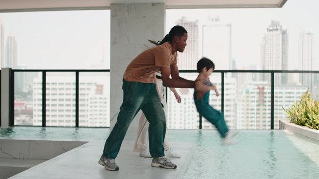 Playful father swinging his son over a rooftop pool as his wife watches
