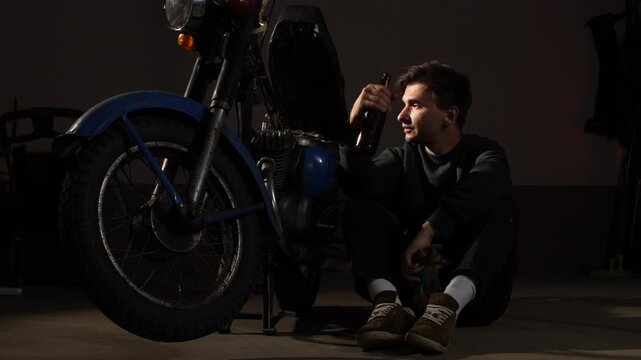 Man sitting on the floor. Holding a beer bottle and a cloth. Resting while attempting to repair a breakdown vintage motorcycle in a dimly lit garage