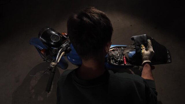 Mechanic working on the open engine of a blue vintage 1970s motorcycle, performing maintenance and repair tasks under dim lighting in a dedicated workspace