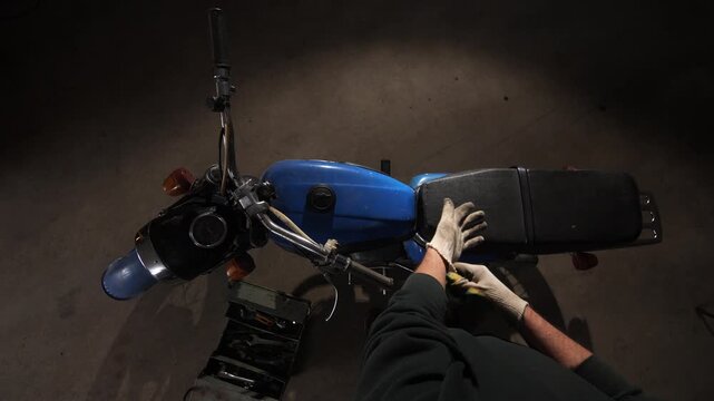 Person wearing work gloves maintaining a classic blue 1970s motorcycle, with a toolbox full of wrenches and tools visible in a dimly lit garage setting