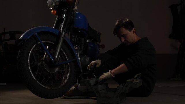 Man in work gloves squatting on the floor, opening a metal toolbox beside a vintage blue motorcycle in a dimly lit garage, performing maintenance
