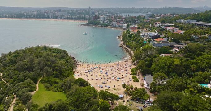 Aerial panorama of Shelly Beach in Australia. People relaxing on the sand, swimming in clear blue water and sunbathing during a warm Australian summer day. Public beach with green park in modern city