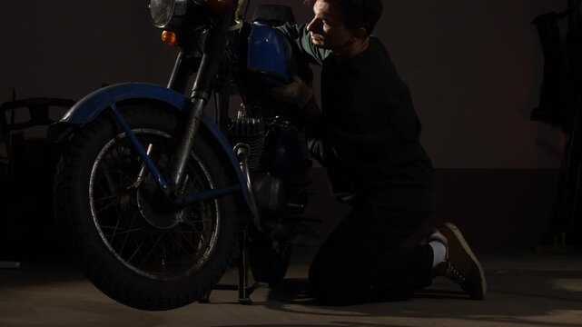 Man kneeling beside a classic 1970s motorcycle, carefully working on its engine with gloved hands, performing maintenance or restoration tasks in a dimly lit garage