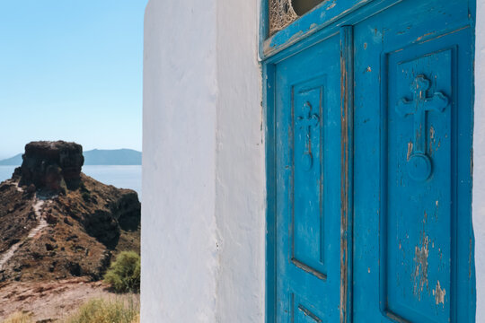 Weathered blue door with carved crosses on a whitewashed wall overlooks a rocky coastal cliff and calm Aegean Sea, capturing the rustic charm and spiritual beauty of traditional Greek architecture.