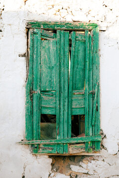 Close‑up of broken green wooden shutters on a whitewashed wall, showing cracked paint and aged texture that reveal the rustic charm and timeless decay of traditional Mediterranean architecture.
