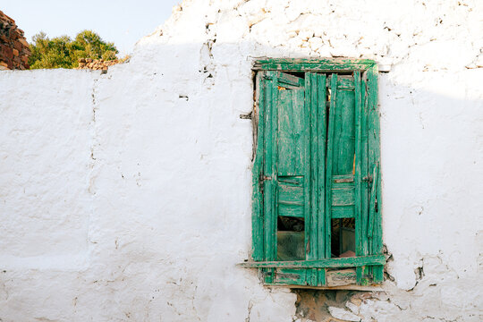 Wide view of a whitewashed wall with broken green shutters and crumbling plaster, capturing the aged beauty and quiet atmosphere of a sunlit Greek island village.