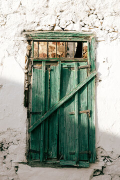 Aged green shutters reinforced with iron bars stand against a cracked white wall, their peeling paint and rough texture evoking the enduring character of historic Mediterranean architecture.