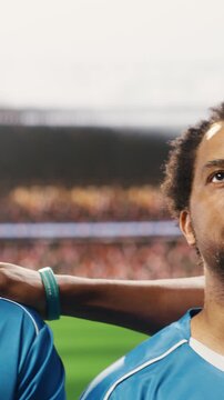 Vertical composition: Diverse soccer players stand together during pre-match anthem ceremony, teammates and goalkeeper in lineup before football game on pitch, blurred stadium crowd in arena.