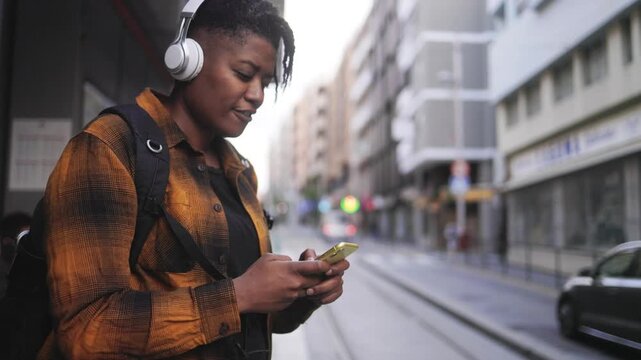 Happy black woman listening to music with mobile phone while waiting at bus station