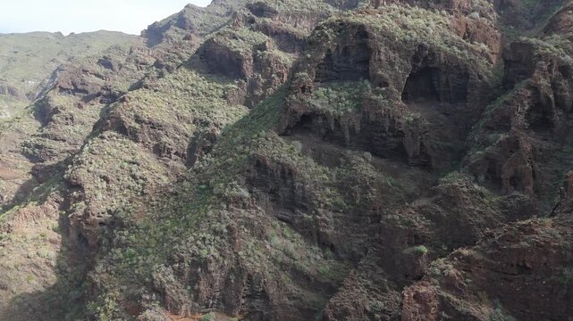Aerial drone footage pushing in and booming up over the rugged, rocky landscape of Mountain Guama on Tenerife. Lush green vegetation clings to the steep cliffs and ridges under a sunny sky.