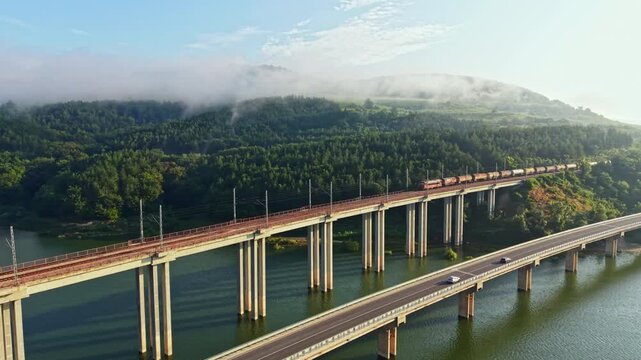 Long concrete bridge spans reflective river while red freight train moves above cars on lower roadway, surrounded by misty forested hills, soft light, calm water and strong perspective