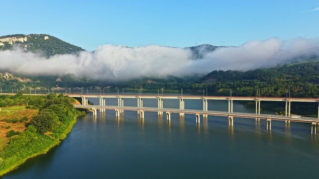 Elevated landscape view showing multi span concrete bridge crossing reflective river while low fog drifts across layered valley hills, with soft morning sunlight warming distant greenery and water
