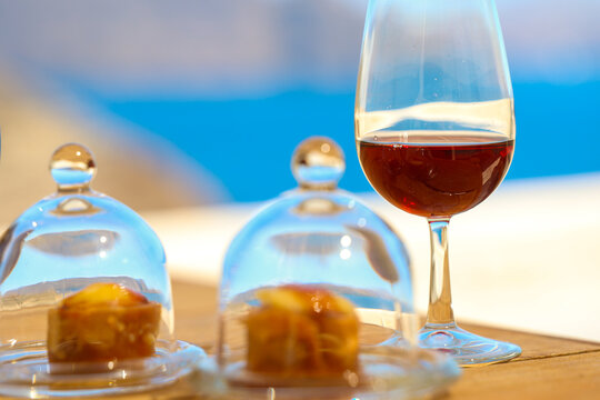 Close‑up of a glass of red wine beside two small pastries covered by glass domes, set against the blurred blue backdrop of the Aegean Sea &mdash; a refined moment of Santorini coastal indulgence.
