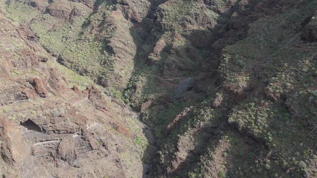 Aerial push in shot of the rugged mountainous landscape of Tenerife. Lush green bushes dot the steep rocky cliffs and deep valley under a sunny sunset sky. A old water canal goes along the cliffs.