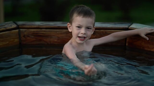 Serious child soaks in wooden outdoor font filled with hot water at twilight, arm extended, peaceful rural setting ideal for childhood relaxation and traditional spa content.
