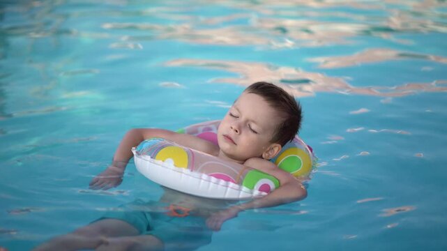 Young boy floats relaxed with eyes closed on colorful inflatable ring in blue outdoor swimming pool during sunny summer vacation.