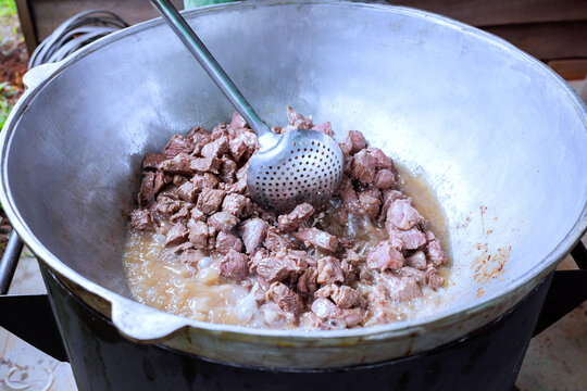 Chunks of meat are being cooked in large pot with broth over an open flame at campsite woods.