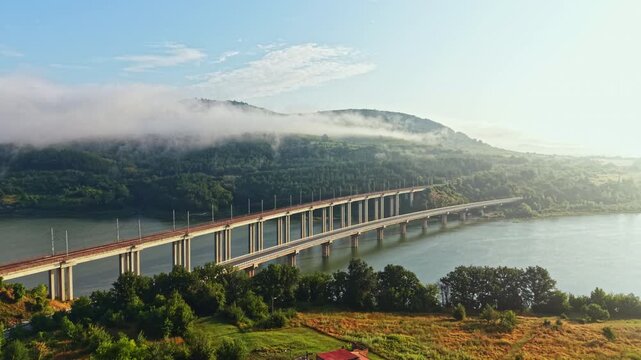 Long girder bridge crosses wide river toward misty hills as fog drifts along water and sunlight glows on deck, creating calm morning scene with reflections, depth, and soft natural atmosphere