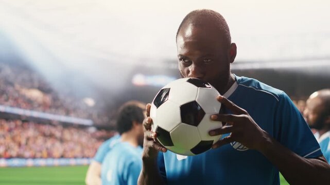 African American soccer player kissing ball on pitch for good luck, wearing blue jersey with teammates blurred behind and stadium crowd in soft focus.