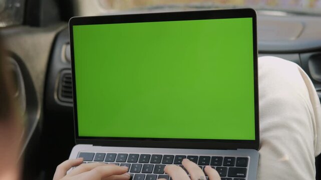 Person types on laptop with green screen inside car. Hands move actively over keyboard. View shows car interior with blurred background. Useful for video editing or remote work scenes