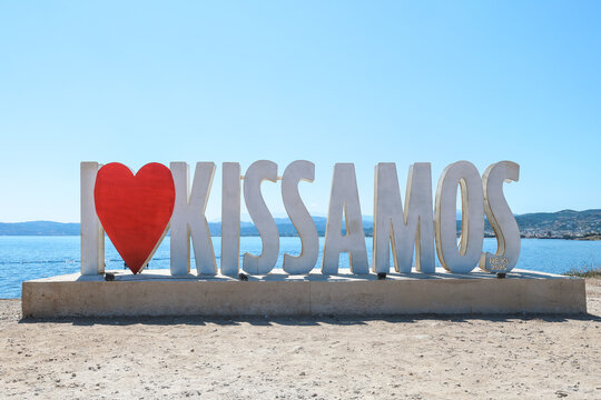 Large "I Love Kissamos" landmark sign on the waterfront in Kissamos, Crete, Greece. Calm Mediterranean waters, distant mountains, and a clear blue sky create an inviting coastal scene.
