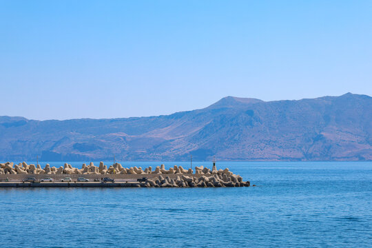 A breakwater extends into blue Mediterranean waters near Kissamos, Crete. Mountains rise across the bay beneath a clear sky, creating a peaceful coastal landscape with maritime character.