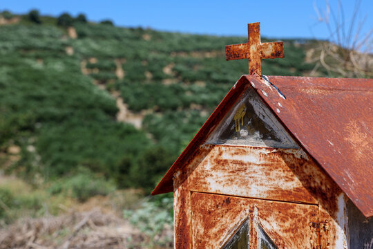 Weathered metal shrine topped with a cross stands in the Cretan countryside. Olive groves cover the hillside behind the structure beneath a clear blue Mediterranean sky.