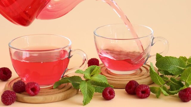 Pouting tasty raspberry tea from teapot into glass cups, fresh berries and mint leaves on beige background, closeup