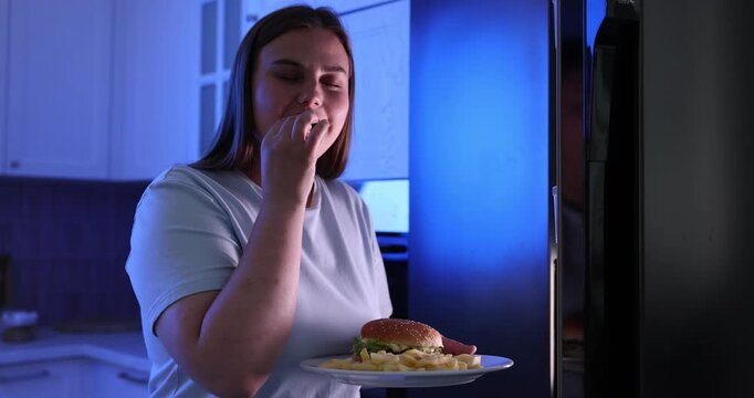 Overweight woman eating French fry from plate with burger near open refrigerator in kitchen at night. Camera moving backwards