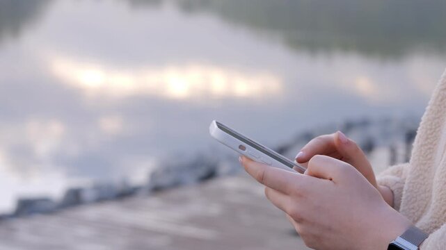 Hand holds smartphone near water's edge. Fingers tap screen, focus on device. Background blurred with distant person. Calm, thoughtful mood. Suitable for social media, travel