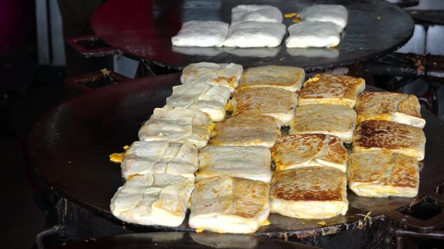 A folded Martabak Telor Kubang pastry filled with seasoned eggs, minced beef, onions, and aromatic spices is carefully placed onto a large hot griddle for cooking. 
