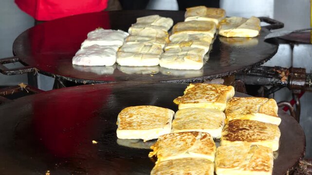 A folded Martabak Telor Kubang pastry filled with seasoned eggs, minced beef, onions, and aromatic spices is carefully placed onto a large hot griddle for cooking. 