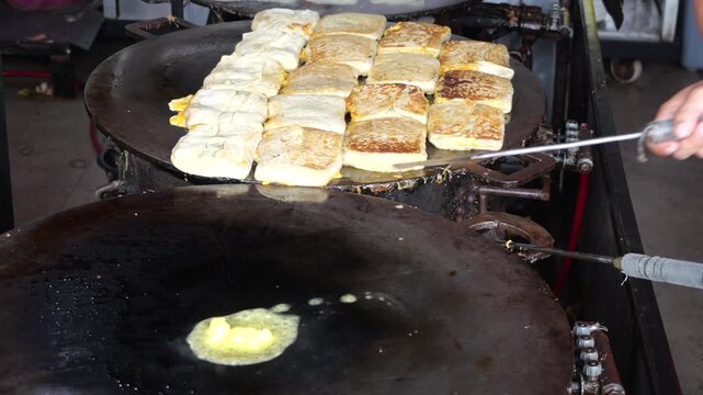 A folded Martabak Telor Kubang pastry filled with seasoned eggs, minced beef, onions, and aromatic spices is carefully placed onto a large hot griddle for cooking. 