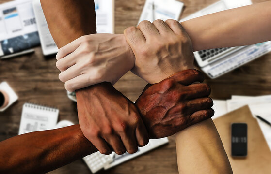 op-view shot of four people holding each other's wrists in a tight circle over a wooden desk with business documents and laptop, symbolizing strength, trust, unity, and strong corporate partnership.