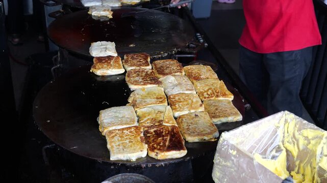 A folded Martabak Telor Kubang pastry filled with seasoned eggs, minced beef, onions, and aromatic spices is carefully placed onto a large hot griddle for cooking. 