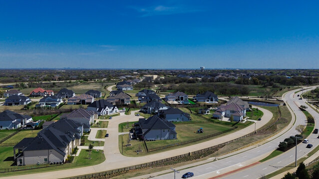 Curved multi lane parkway borders Dallas suburb Tour 18 subdivision. Wide aerial shot displays executive custom homes, cul de sacs, distant water tower. Flower Mound, TX