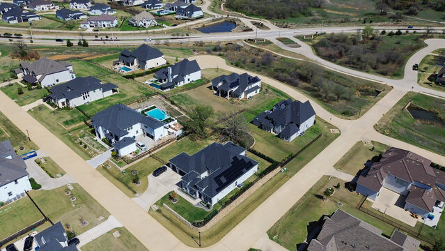 Solar panels cover modern dark rooflines inside Dallas suburb Chimney Rock neighborhood. High altitude view depicts paved access roads, swimming pools, lush lawns, open ground. Flower Mound, TX