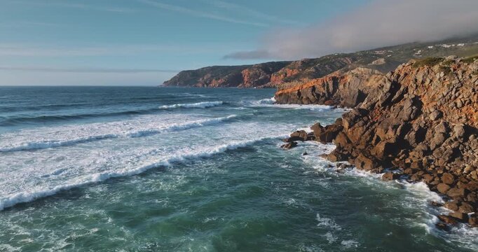 Rolling ocean waves crashing against rugged coastal cliffs and a rocky shoreline, showcasing the natural beauty of Sintra Cascais Natural Park in Portugal under a clear blue sky. Natural background